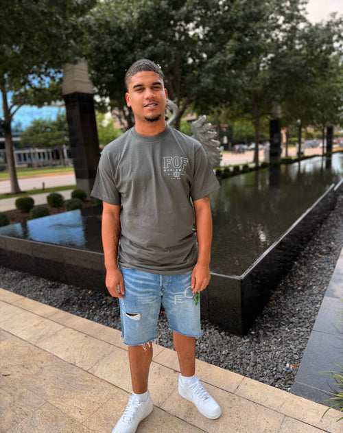 A man standing outdoors in front of a water feature, wearing a gray t-shirt with a white graphic, distressed denim shorts, and white sneakers.